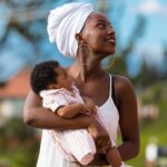 A joyful African mother holding her baby under sunny blue skies outdoors.